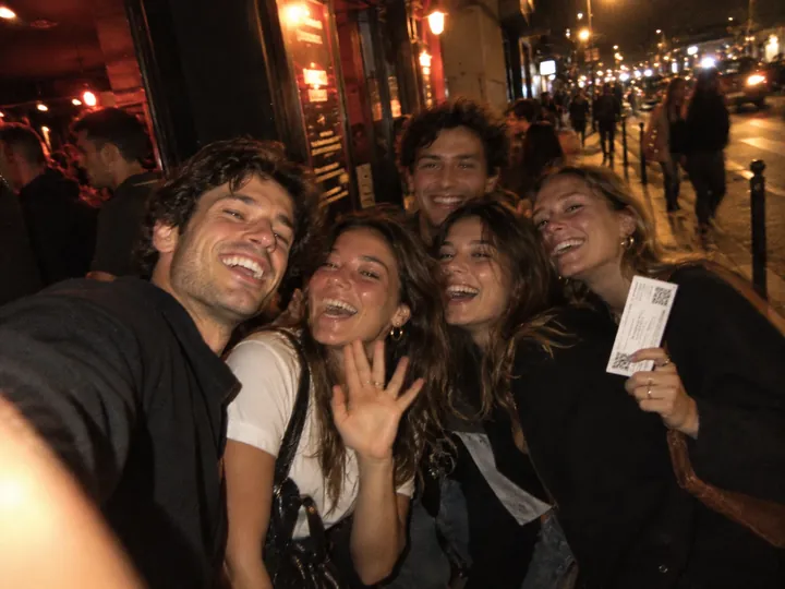 A man taking a selfie with laughing followers outside a venue at night, one holding up her ticket